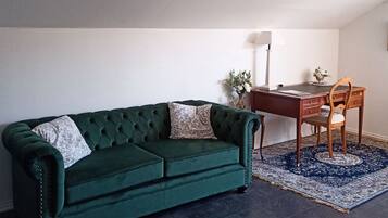 Un sofá de terciopelo verde con dos almohadones, un escritorio de madera, una silla, una lámpara y una alfombra estampada en una habitación con techo inclinado.