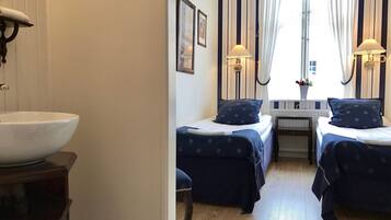 Quarto de hotel com uma pia grande branca, duas camas com roupa de cama azul combinando, uma mesa pequena e uma janela com cortinas.
