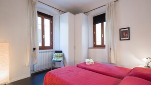 A room with two beds, a radiator, a chair, and a window with curtains.