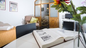 A hotel room with a bed, a desk with an open book, a chair, a small table, and a decorative plant.