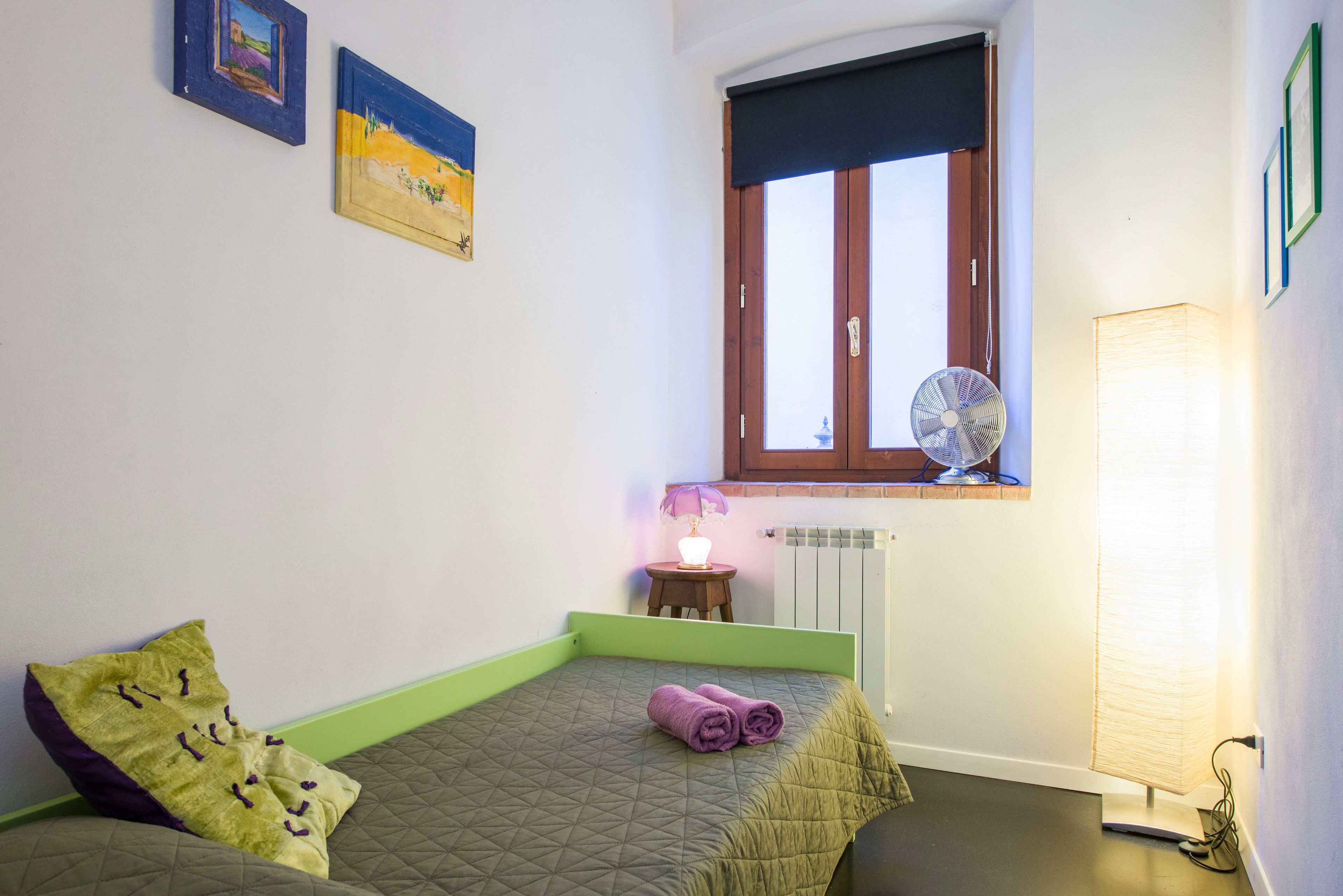 A small bedroom with a bed, a window, a fan, and a lamp.