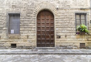 Ein Steinhaus mit einer großen, verzierten Holztür, einem kleinen Fenster mit Metallgitter und einem Blumentopf auf der rechten Seite.