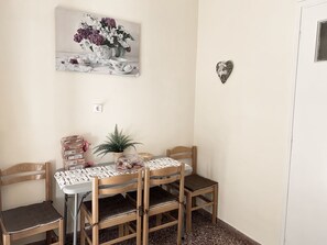 Un coin repas avec une table dressée pour quatre personnes, entourée de chaises en bois avec des coussins. Une plante en pot et un objet décoratif portant le mot « MAISON » sont posés sur la table.