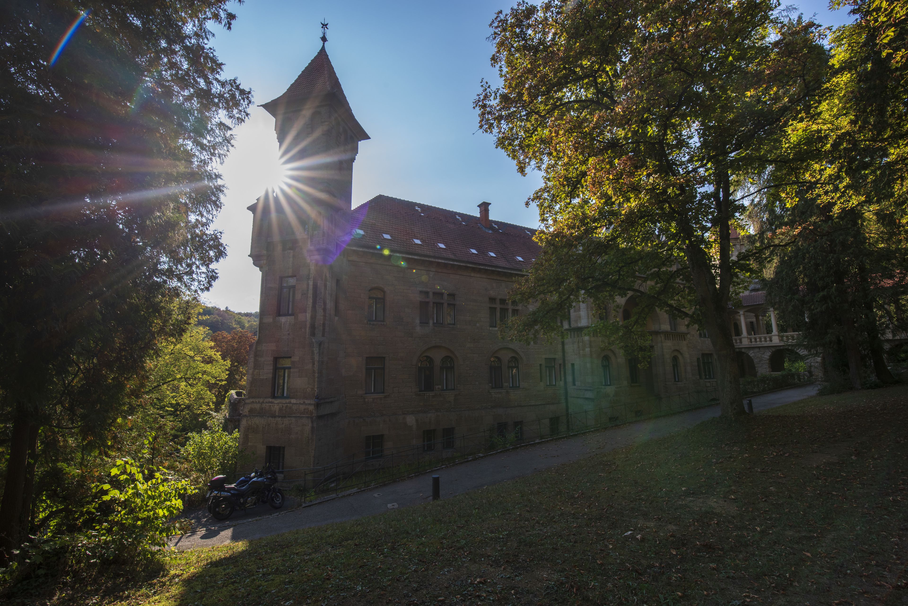 A historic building with a prominent tower and arched windows, surrounded by trees and a clear sky.