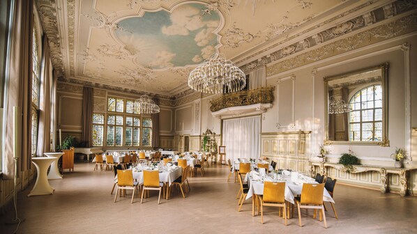 A grand dining hall with ornate ceiling and chandeliers, large windows, and formal table settings.