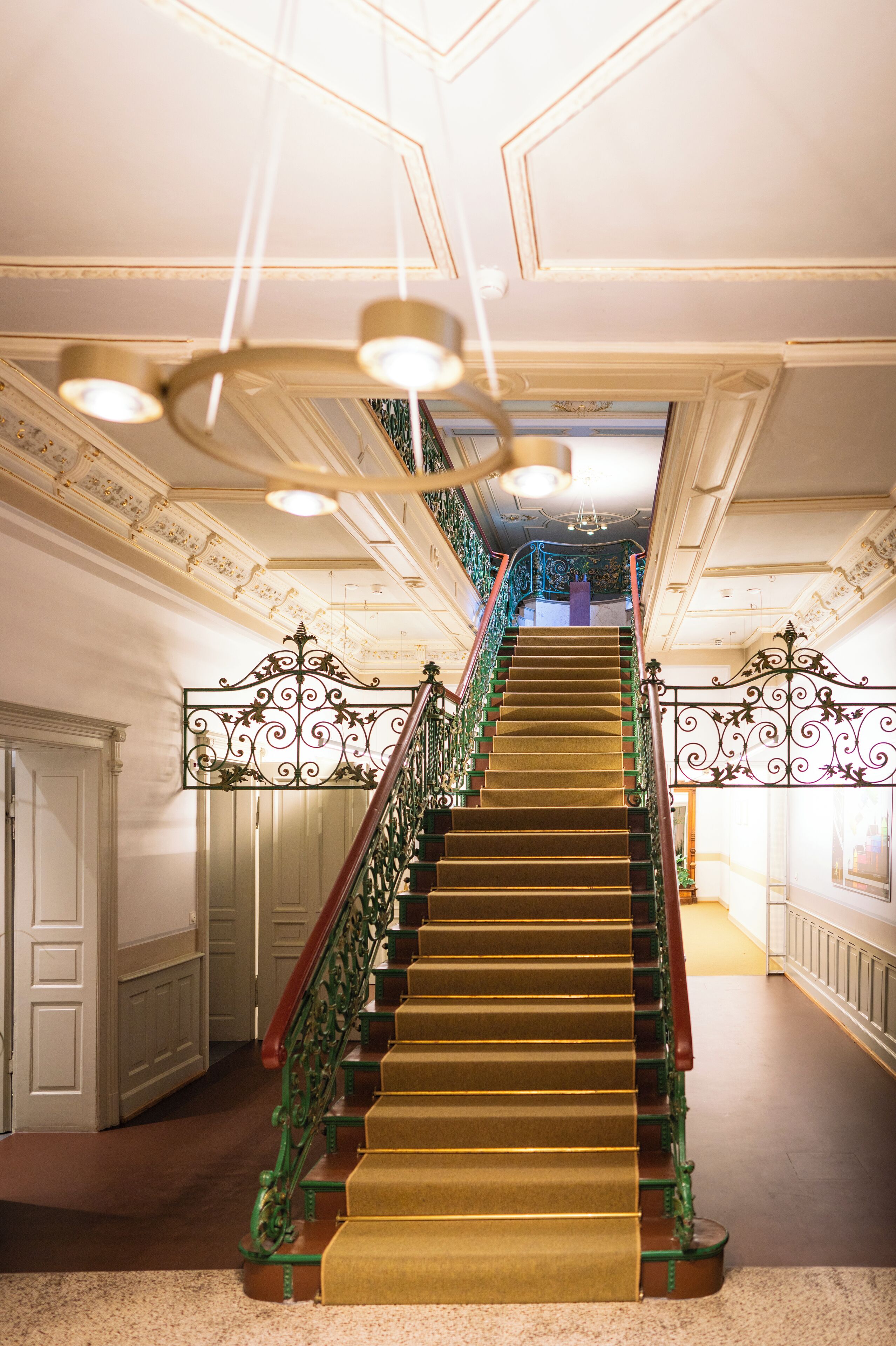 A grand staircase with ornate metal railings and a carpeted floor, leading to a landing with a view of the interior.