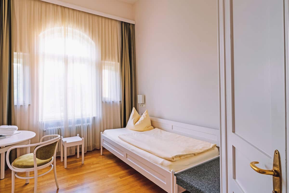 A single bed with a white headboard, a bedside table, a chair, and a window with curtains.