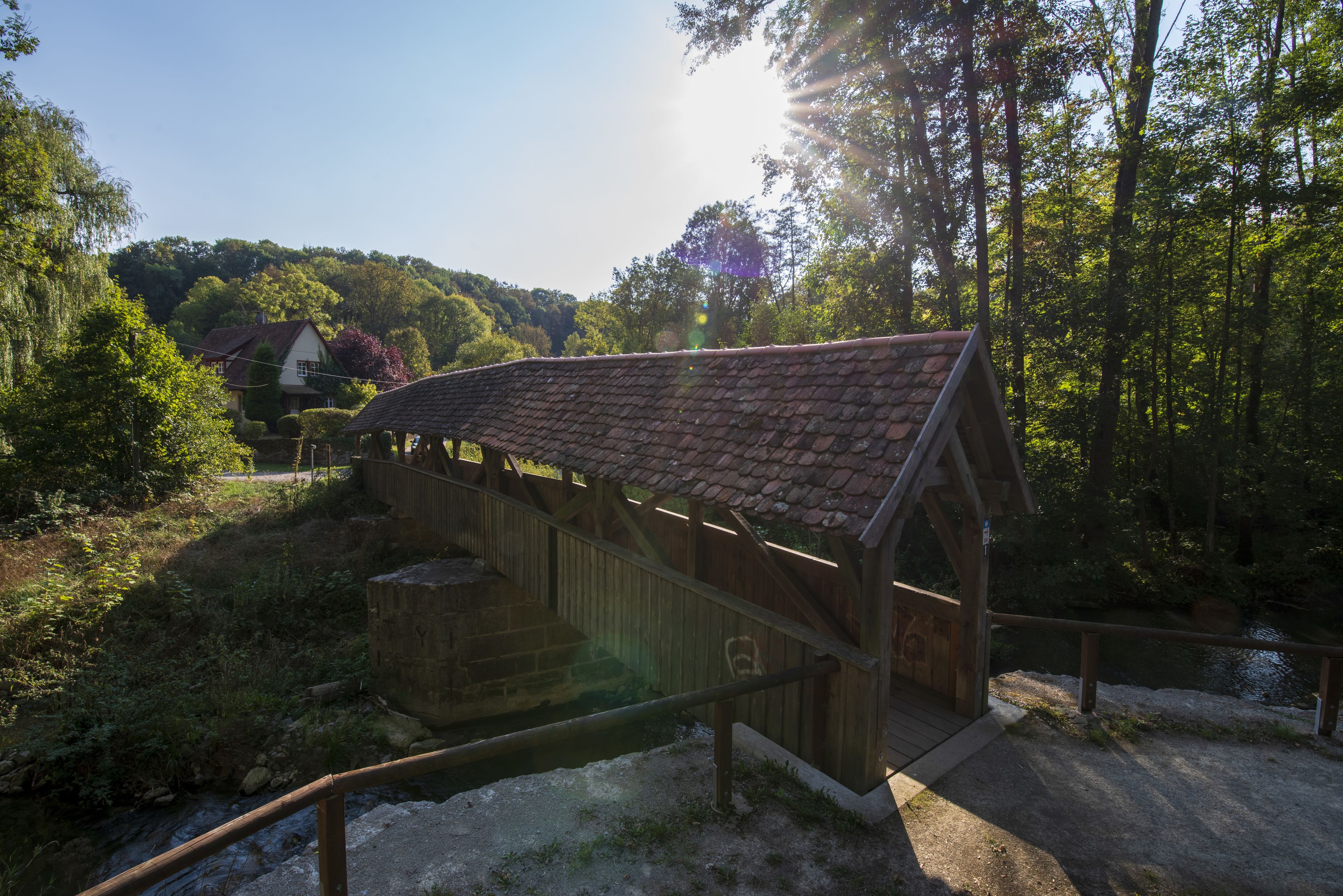 Eine hölzerne gedeckte Brücke mit Metallgeländer, umgeben von viel Grün und blauem Himmel.