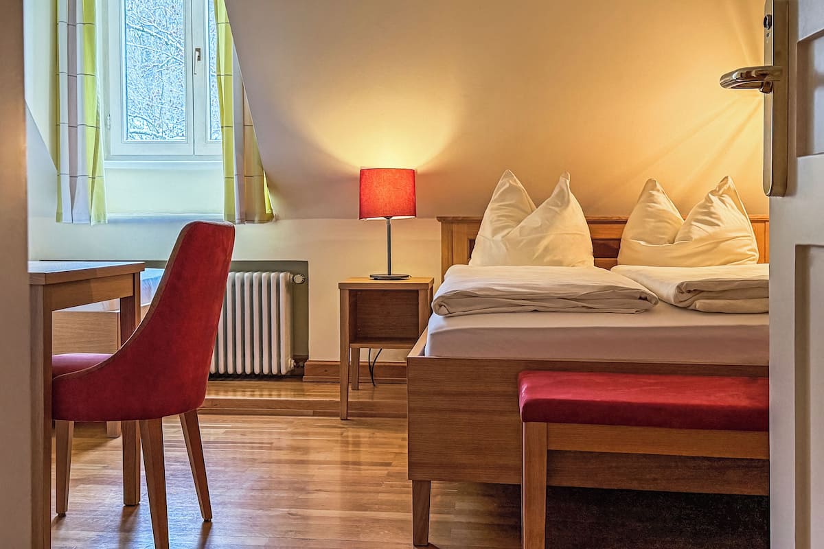 A hotel room with a bed, a desk, a chair, a lamp, and a window.