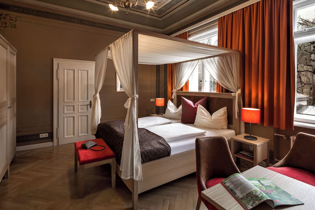 A four-poster canopy bed with a bedside table, a reading chair, and a small desk with a lamp.