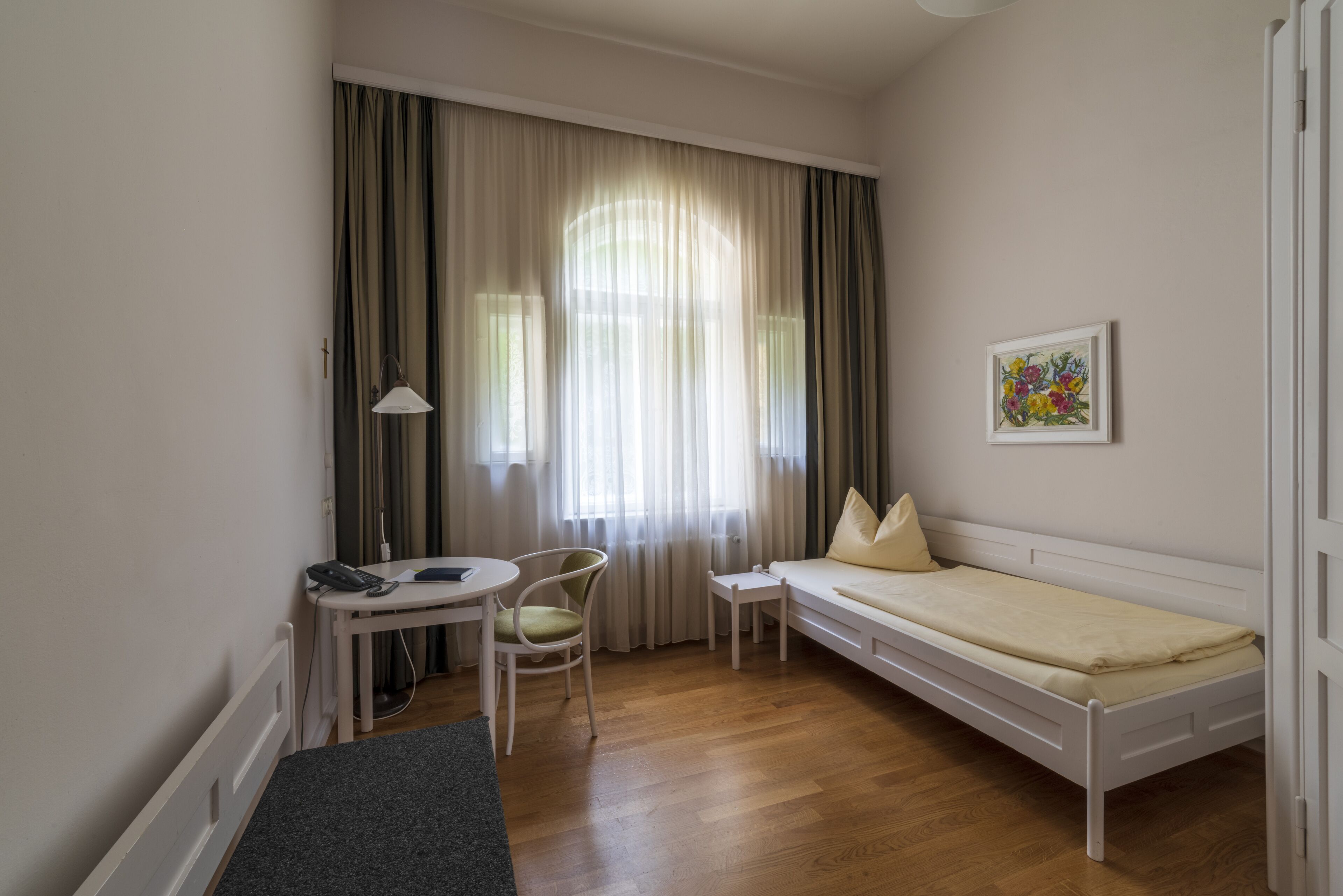 Quarto pequeno e bem iluminado com uma cama, escrivaninha, cadeira e janela com cortinas.