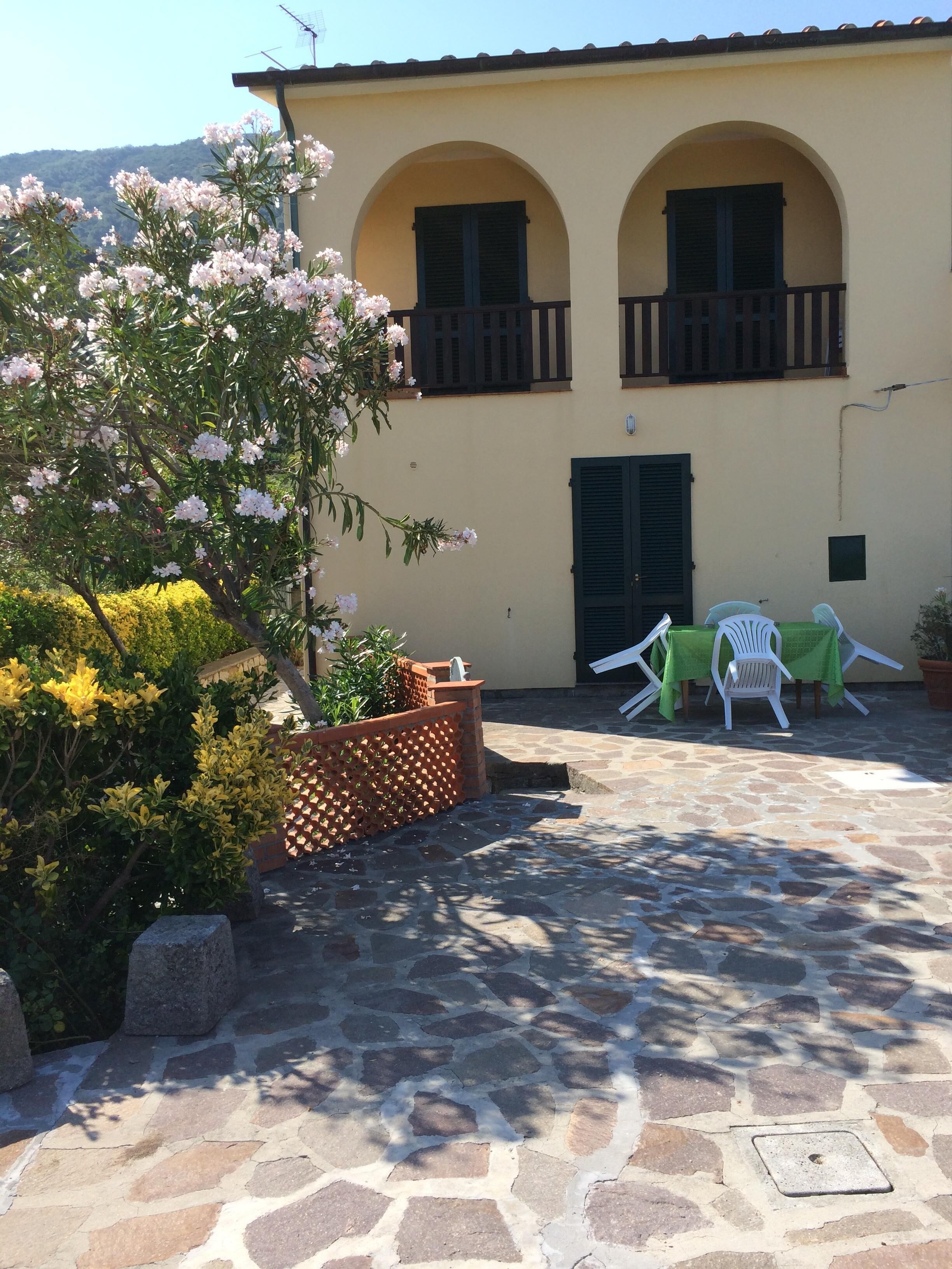 A courtyard with a stone floor, a building with arched windows and a balcony, and a table with chairs set up outside.