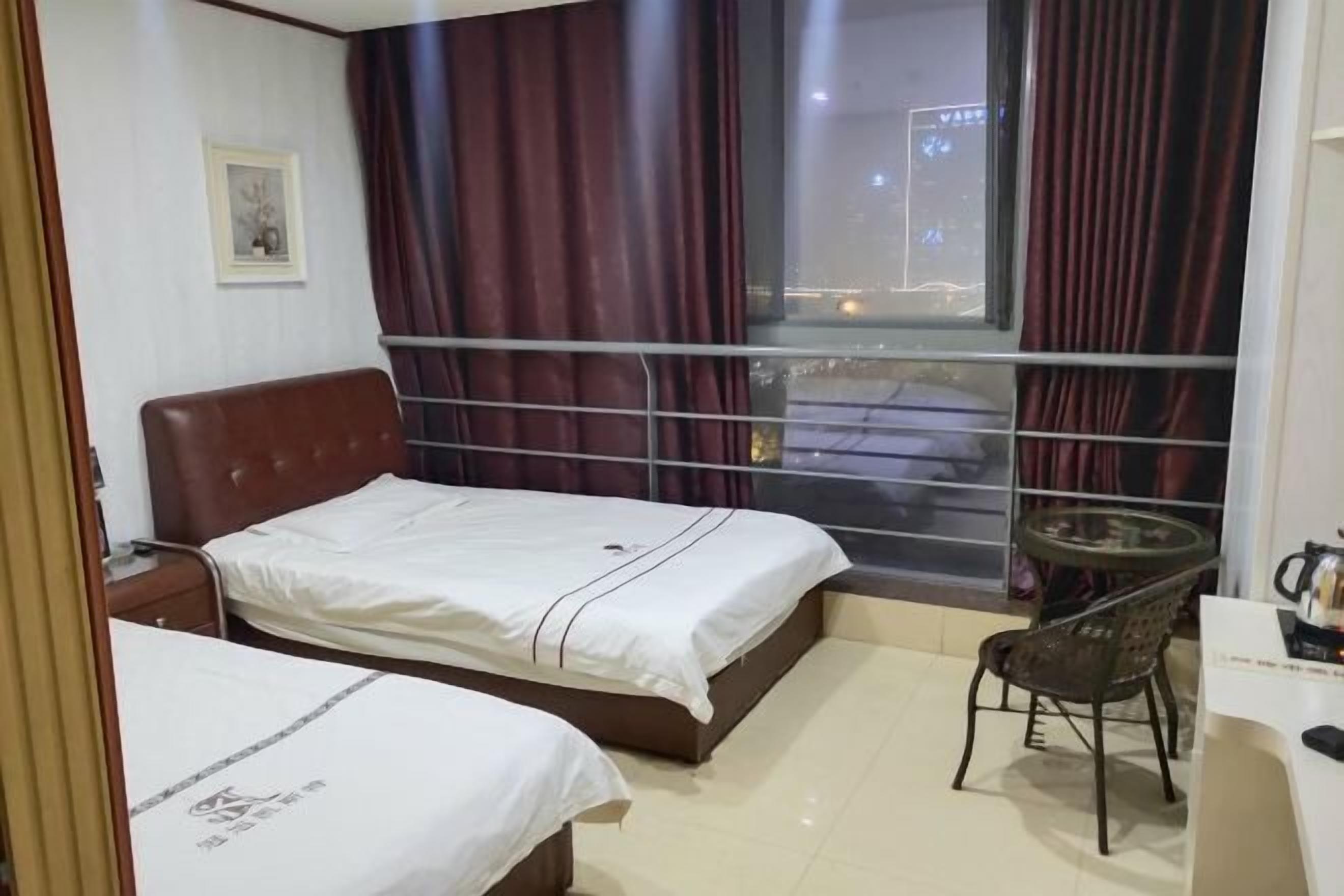 A hotel room with two beds, a small table with a chair, a window with curtains, and a framed picture on the wall.
