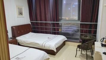 A hotel room with two beds, a small table with a chair, a window with curtains, and a framed picture on the wall.