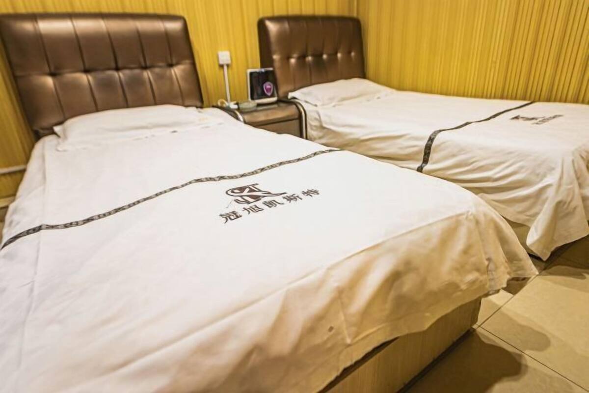 Een hotelkamer met twee bedden, elk voorzien van een witte spreidek en een logo. Er is een nachtkastje met een lamp en een klein tafeltje met een decoratief object.