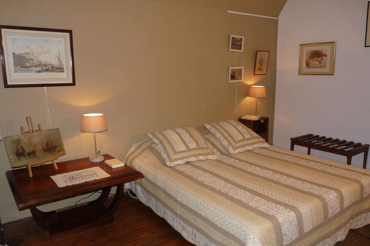 Un dormitorio ordenado con cama, mesitas de noche, un escritorio con lámpara y cuadros en las paredes.