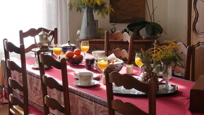 Una mesa de comedor con mantel rojo, sillas de madera, un florero con flores amarillas y diversos alimentos para el desayuno, como jugo de naranja, pasteles y una tetera.