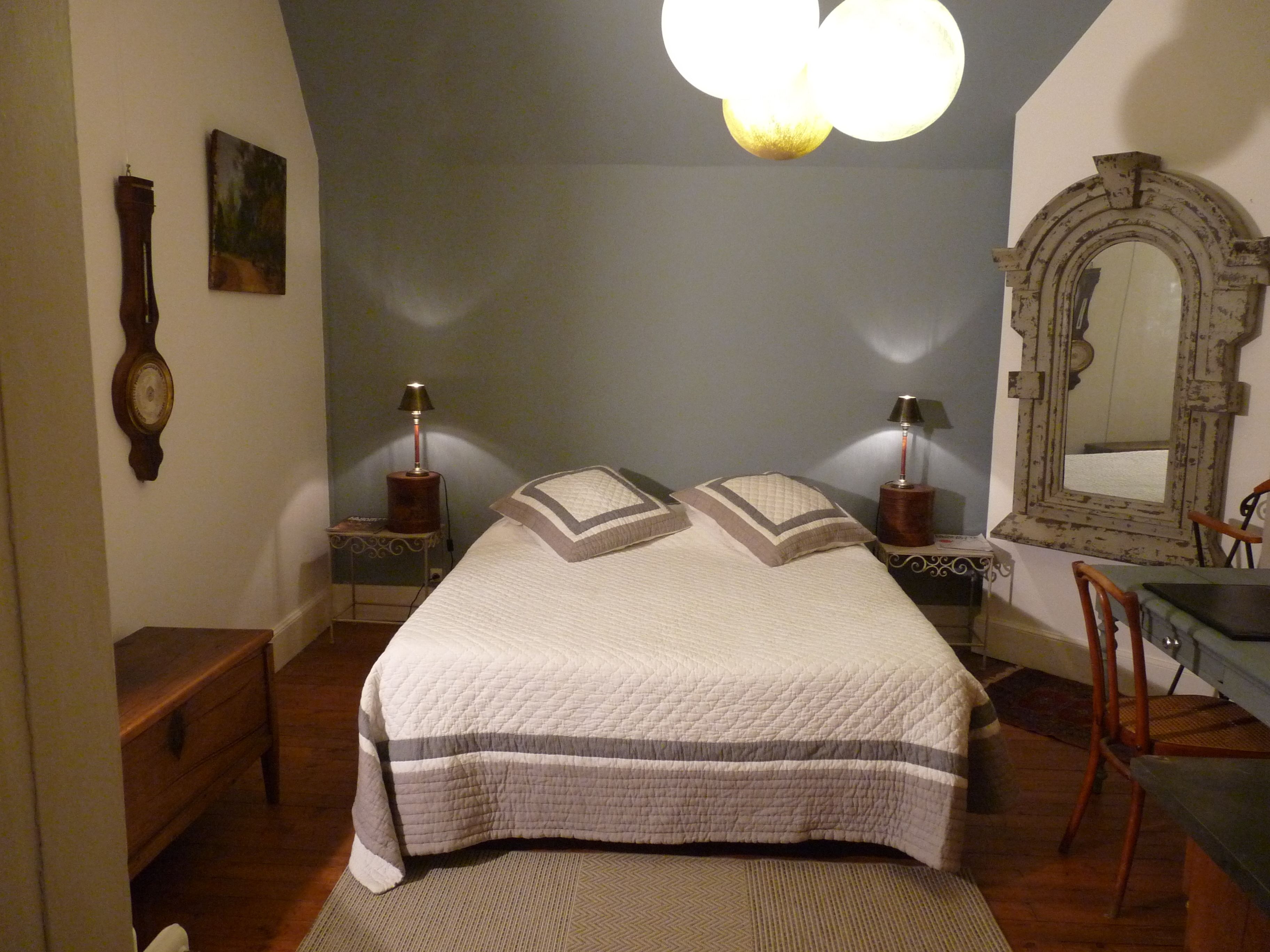 Ein Schlafzimmer mit einem großen Bett, zwei Nachttischen mit Lampen, einem Holzstuhl, einem Schreibtisch, einem Spiegel und einer Wanduhr.