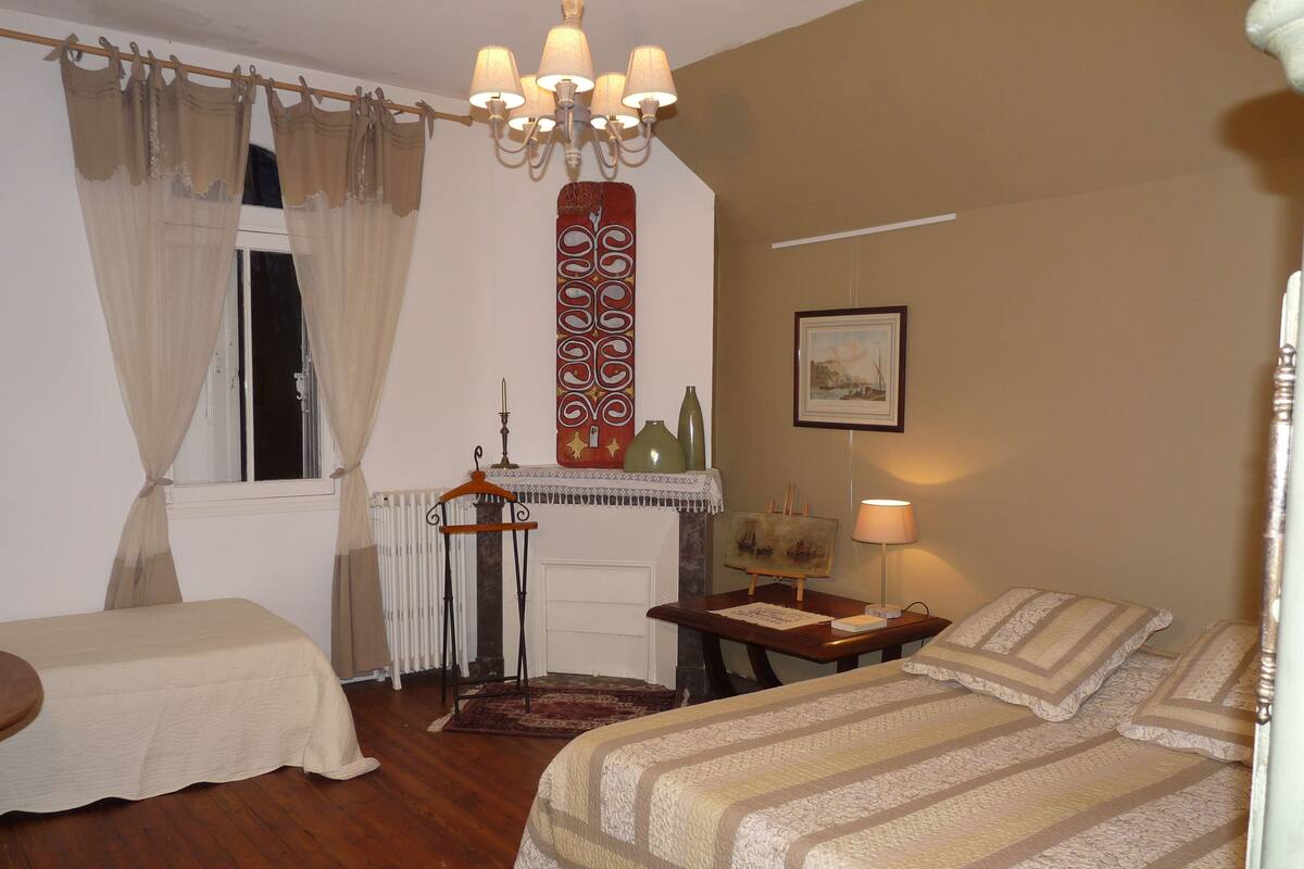 Un dormitorio con dos camas, un aparador, una mesita de noche, una ventana con cortinas, un candelabro, un cuadro y una lámpara.