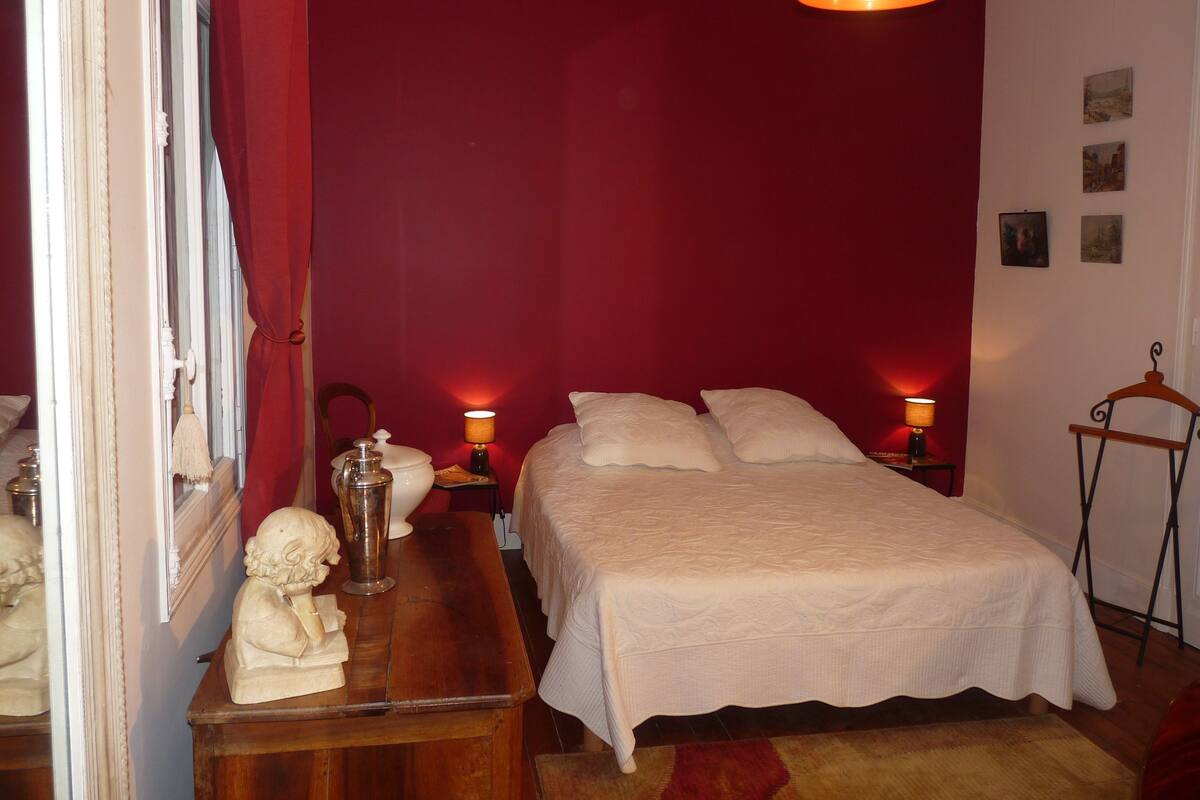 Un dormitorio con cama, lámparas de noche, una mesita de noche de madera, pared roja y una lámpara colgante.