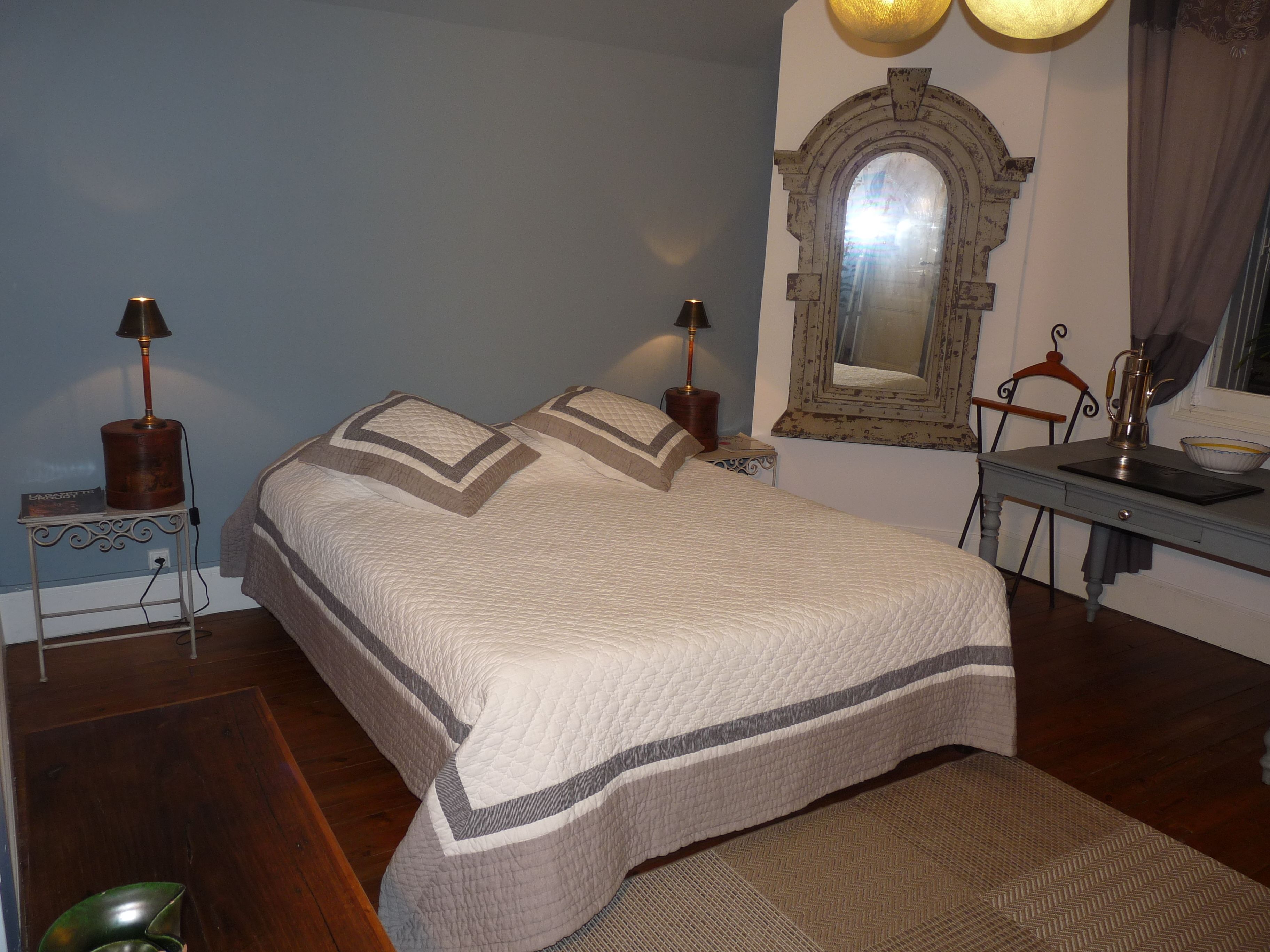 Una cama bien hecha con colcha, dos lámparas de noche, una mesita con lámpara, un espejo y una silla.