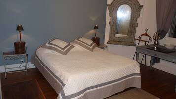Una cama bien hecha con colcha, dos lámparas de noche, una mesita con lámpara, un espejo y una silla.