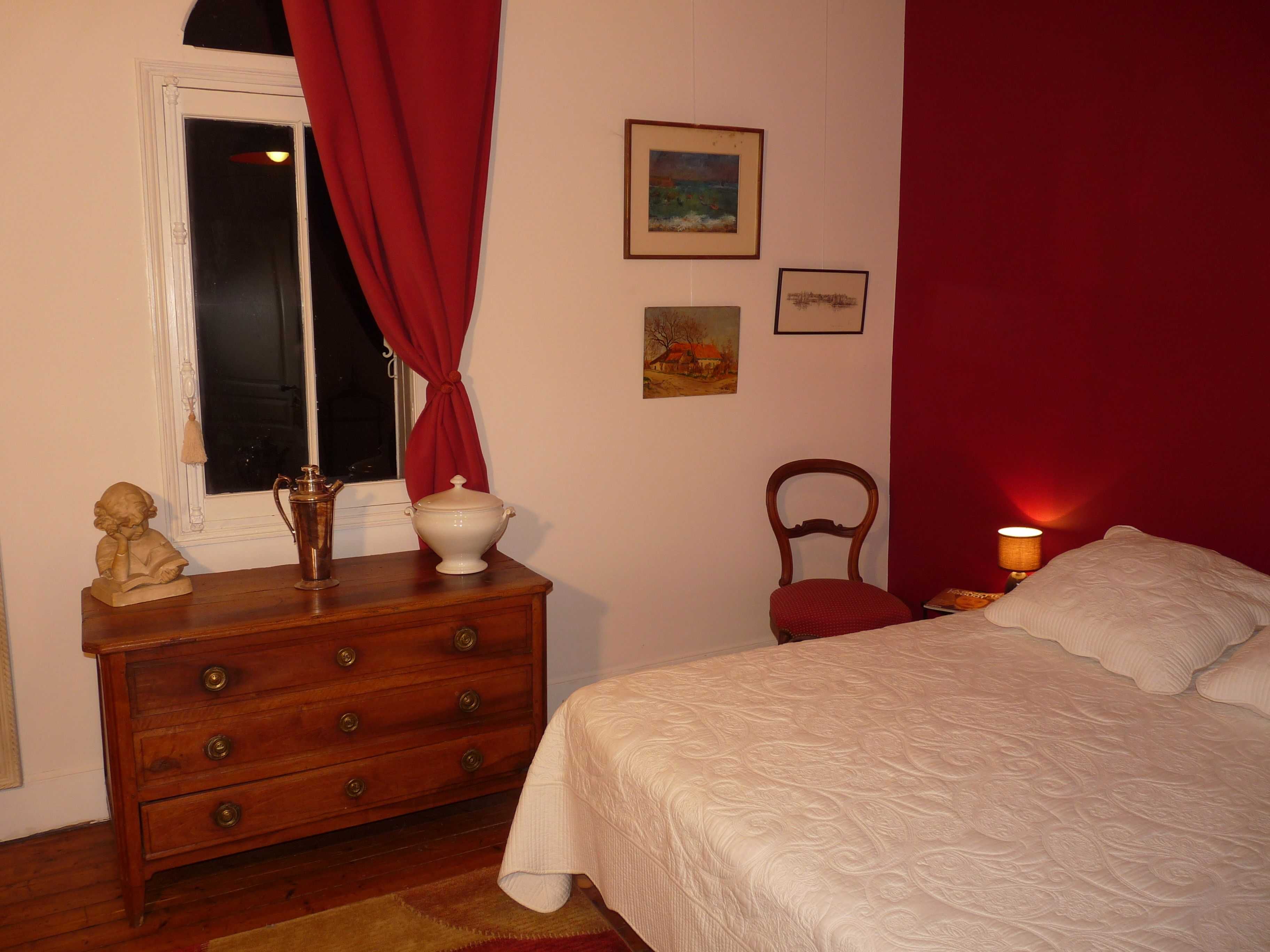 Un dormitorio con un aparador de madera, una cama con ropa blanca, una silla roja y una mesita de noche con una lámpara.