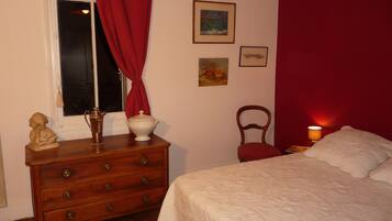 Un dormitorio con un aparador de madera, una cama con ropa blanca, una silla roja y una mesita de noche con una lámpara.