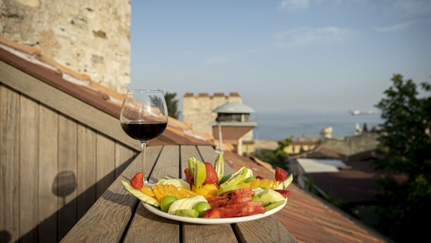 Une table en bois, sur laquelle se trouvent une assiette de fruits et un verre de vin rouge, avec vue sur la côte, des bâtiments et un mur de pierre.