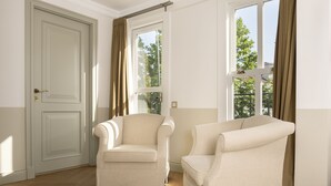 Une pièce avec deux fauteuils beiges, un plancher en bois, une porte blanche et des fenêtres avec des rideaux.