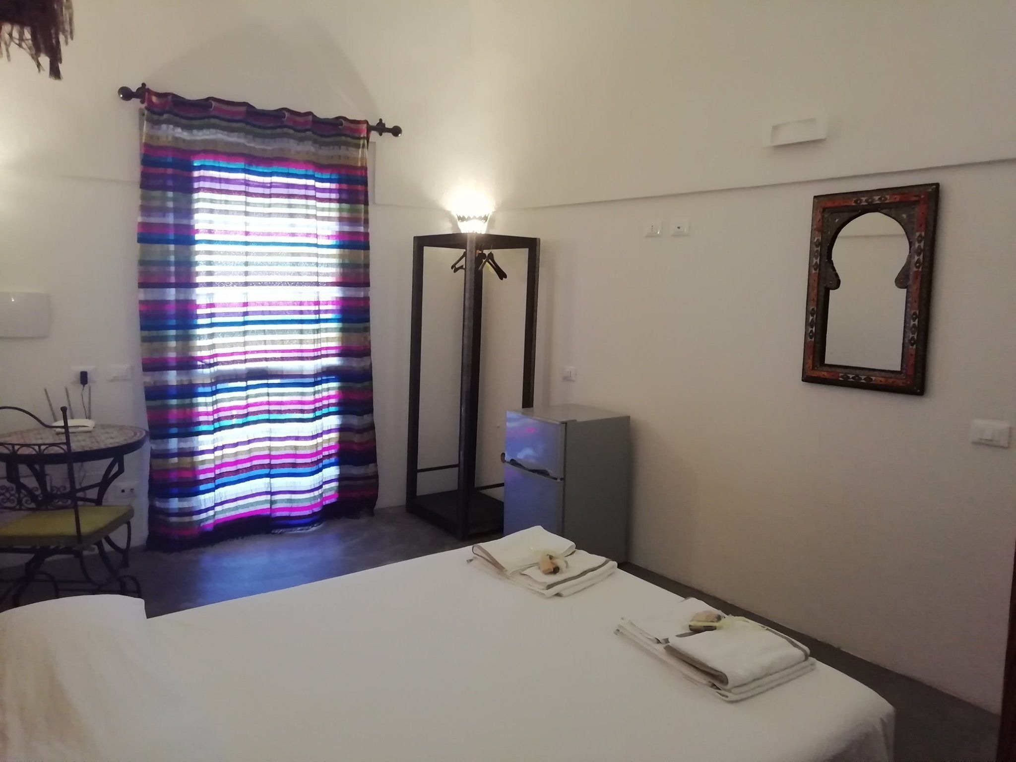 A hotel room with a bed, a bedside table, a chair, a mirror, and a curtain.