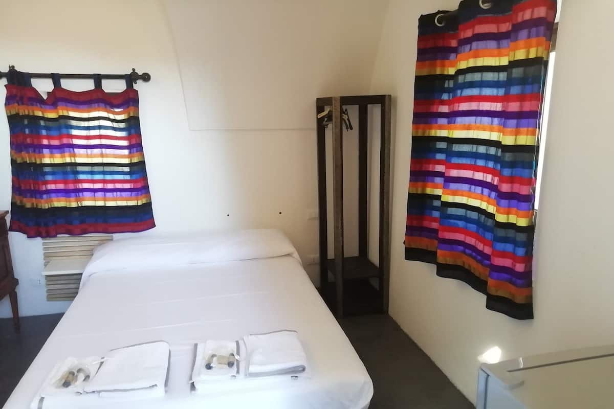 Une chambre d’hôtel avec un lit, un banc en bois, une petite table et un étagère en métal.