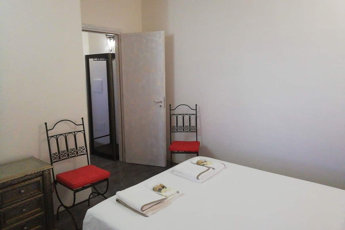 Une petite pièce simple, avec un lit, deux chaises, une petite table et une porte donnant sur une autre pièce.