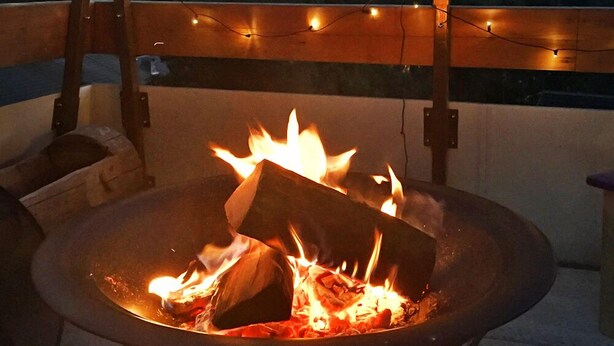 Una hoguera con leña ardiendo, rodeada de un banco de madera y luces colgantes, con montañas al fondo.