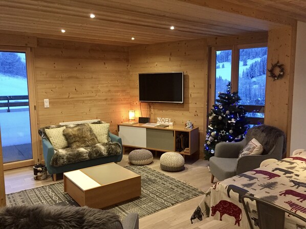 Un salon chaleureux avec un décor intérieur en bois, un téléviseur à écran plat fixé au mur, un sapin de Noël et un canapé agrémenté d’un plaid en fourrure.