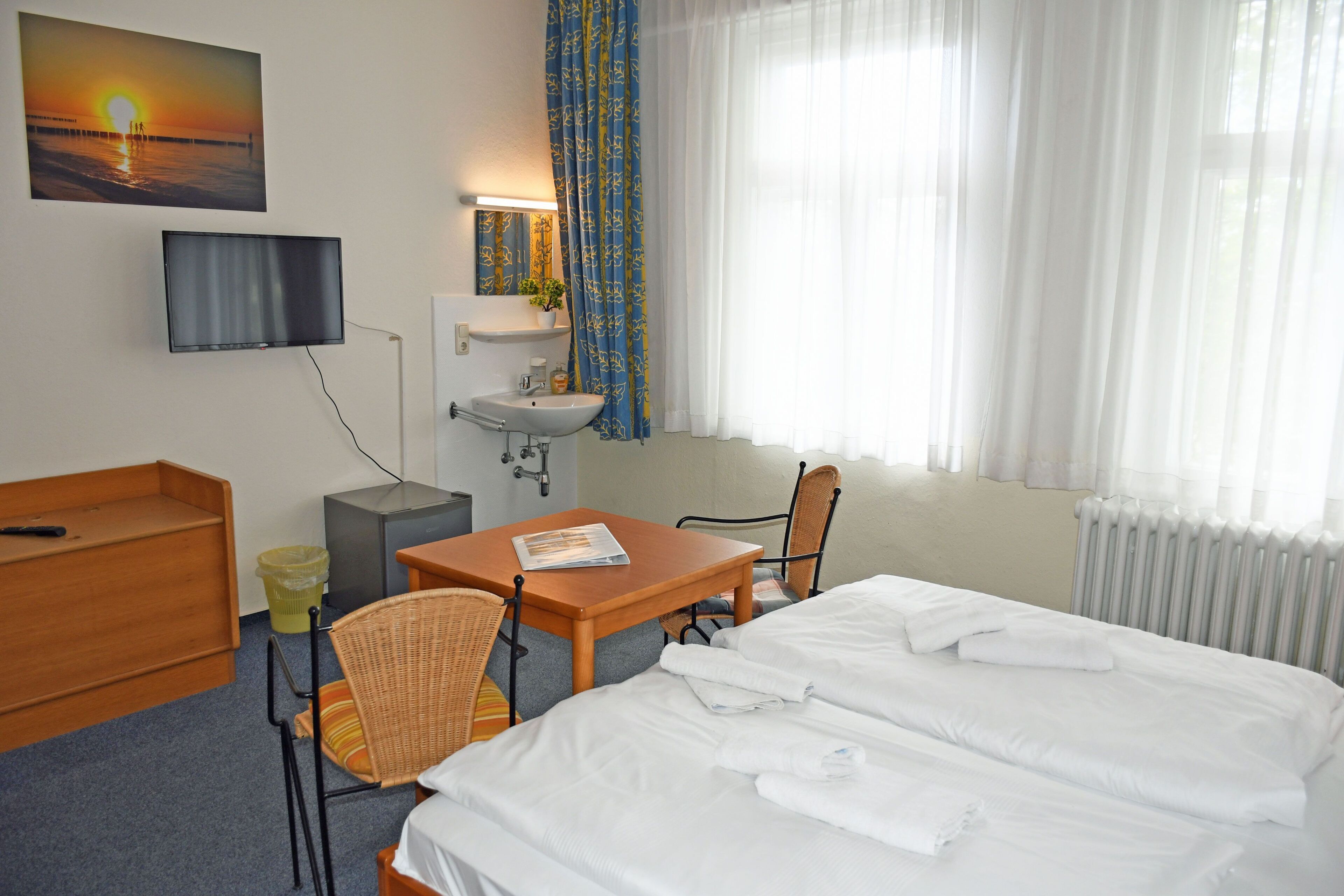 A hotel room with a bed, a desk, a chair, a TV, a sink, and a window with curtains.