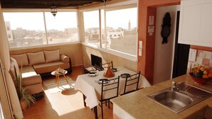 A cozy dining area with a table set for two, a sofa with patterned cushions, and a view of the cityscape through large windows.