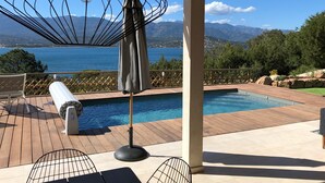 Un espace aménagé au bord de la piscine, offrant une vue sur la mer et les montagnes, avec une terrasse en bois, des chaises longues et un parasol.