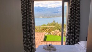 Une chambre dotée d’un grand lit, de draps et de serviettes blancs, d’une terrasse en bois offrant une vue sur un lac et des montagnes, et de rideaux à motifs.