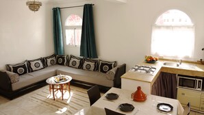 Ein Wohnzimmer mit einer Couch, einem Esstisch für zwei Personen, einer Küchenzeile mit Herd und Mikrowelle sowie einem Fenster mit Vorhängen.