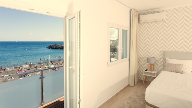 A hotel room with a large window overlooking a beach, a bed, a bedside table with a lamp, and a view of the sea. - Beach House by Azores Villas (Ponta Delgada)