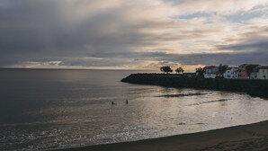 A coastal scene with a calm sea, a rocky breakwater, and buildings along the shore. - Beach House by Azores Villas (Ponta Delgada)