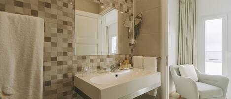 A modern bathroom with a checkered tile backsplash, a large mirror, a white sink, and a light-colored armchair in the adjacent room.