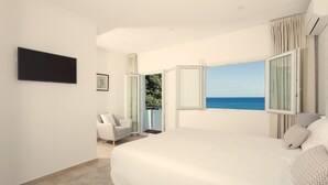 A bedroom with a large bed, a TV mounted on the wall, a chair, and a view of the sea through open windows. - Beach House by Azores Villas (Ponta Delgada)