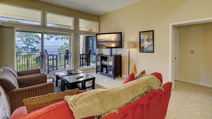 Un soggiorno con un divano rosso, un tavolino in vetro e una TV che mostra 'VACATION INCOMPANY'.  C'è un balcone con vista sul mare.