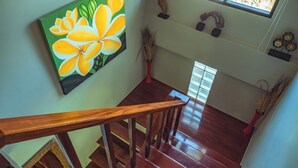 A wooden staircase with a painting of yellow flowers on the wall, a red vase, and a window with a view of greenery.
