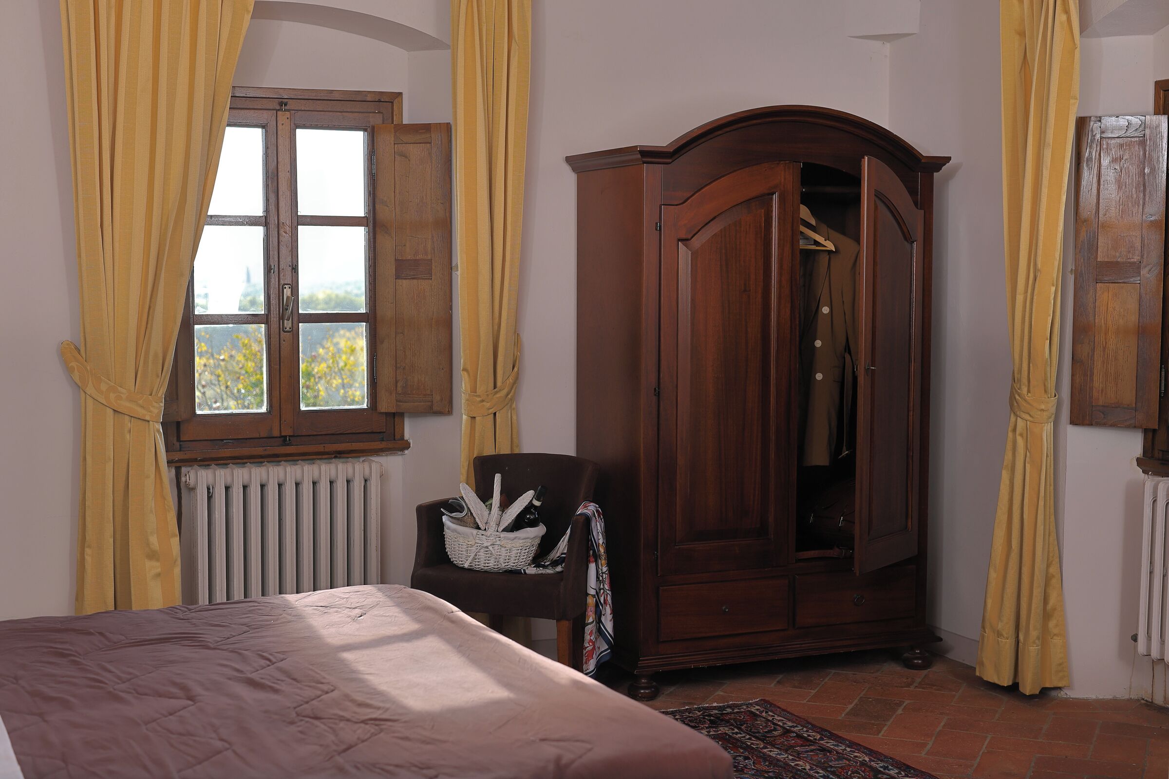 Une chambre à coucher avec un lit, une armoire en bois, une chaise avec un panier, et une fenêtre avec des rideaux.