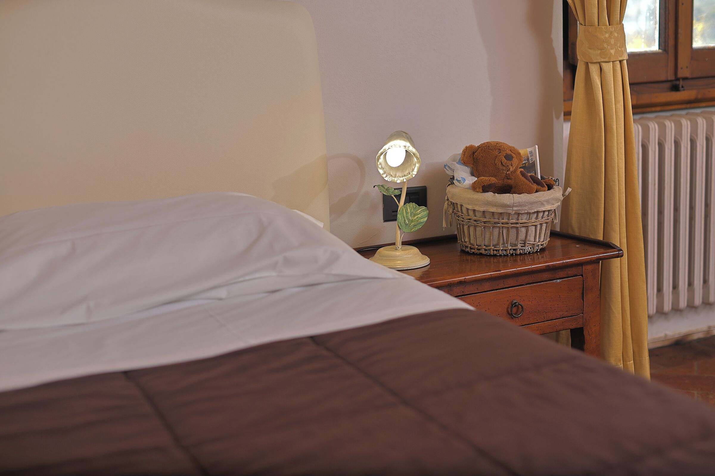 Een net opgemaakt bed met witte lakens, een nachtkastje met een lamp, een teddybeer en een mandje.