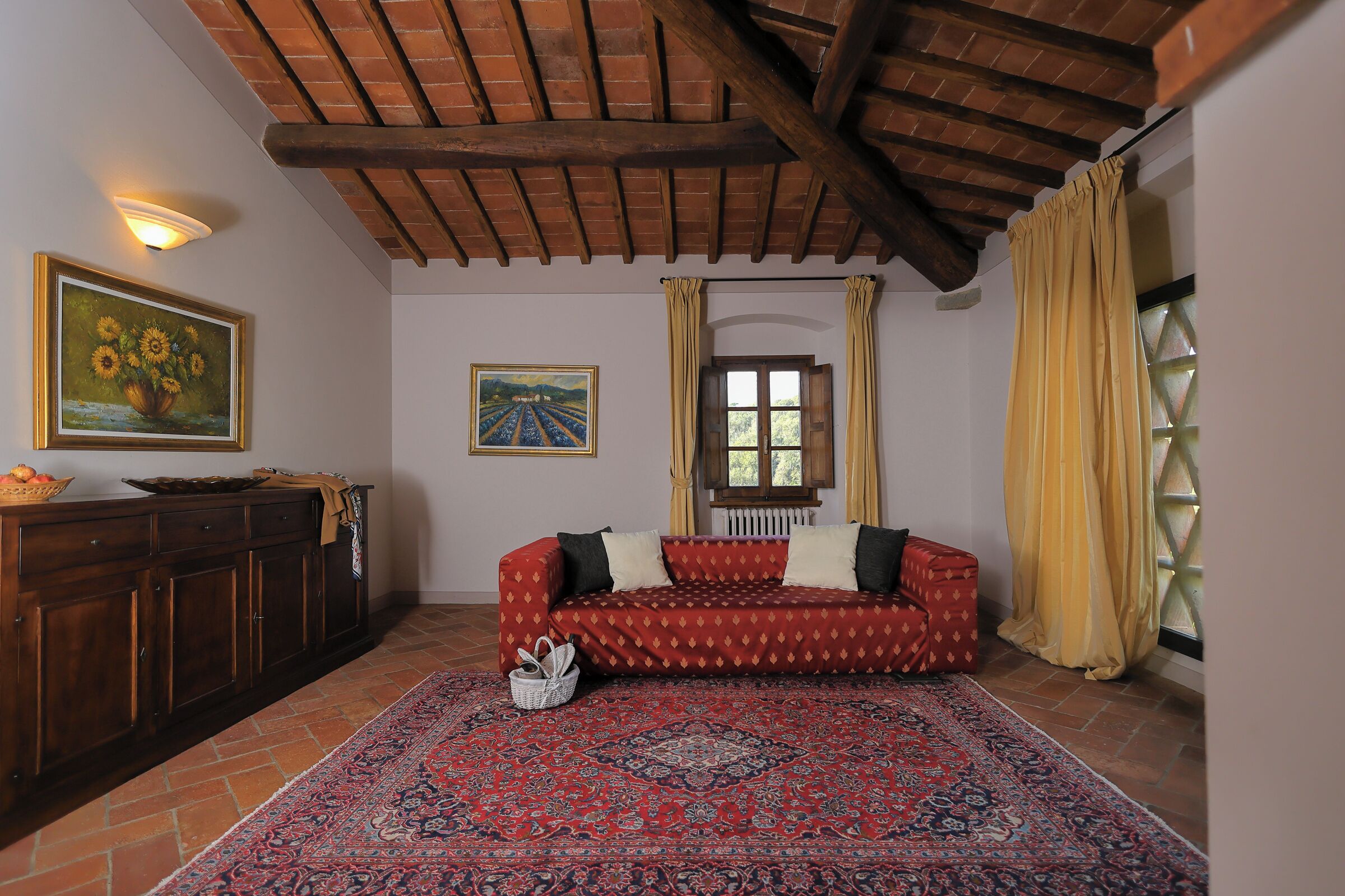A cozy living room with a patterned rug, a wooden ceiling, a sofa, and a window with curtains.