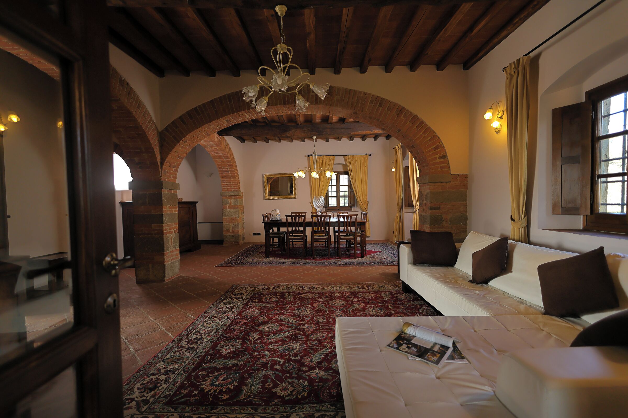 A spacious living area with a high ceiling, exposed wooden beams, a dining table with chairs, a white sofa with cushions, and a patterned rug.